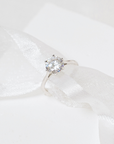 Ella round solitaire ring with a slim silver-tone band, resting on folded white satin.