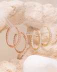 Holly hoops with round clear stones in rose-gold and yellow-gold tones, hanging from white mesh on pale stone.