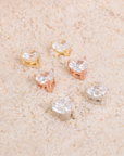 Penelope heart-shaped clear stud earrings in silver, yellow-gold and rose-gold tones on pale stone.